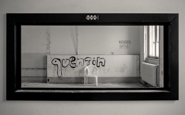 Schwarz-Weiß Bild im Landscape Format. Zu sehen ist ein breiter Schwarzer Rahmen von einem ehemaligem einseitigen Spiegel dem jetzt aber die Scheibe fehlt. Dahinter liegt wie gerahmt der ehemalige Vernehmungsraum. Dieser ist allerdings quasi leer nur ein weißer, leerer Gartenstuhl steht in der Bildmitte. Dahinter steht als Graffiti quentin. Rechts im Bild sind ein Heizkörper und ein Fenster teilweise zu sehen.