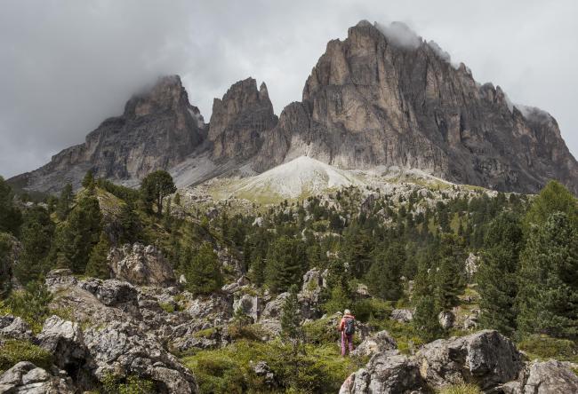 Farbbild im Landscape Format. Das Bild zeigt ebenfalls das Langkofelmassiv aus der Steinernen Stadt heraus aber in einem deutlich weiteren Winkel. Im Vordergrund bis in die Bildmitte sind verschiedene Große Felsen in der Landschaft zu sehen. Dazwischen und richtung Bildmitte folgen dann Nadelbäume. Dahinter folgt das Langkofelmassiv mit einem Bedeckten Himmel und ein paar Wolken an der Bergspitze. Vorne im Vordergrund etwa leicht rechts der Mitte steht eine Person mit roter Kleidung und schaut den Berg hinauf.