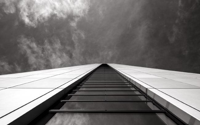 Schwarzweißbild im Landscape Format. Die obere Hälfte des Bildes besteht aus Himmel mit ein paar hellen sehr feinen Wolken. Die untere Hälfte des Bildes zeigt eine Fassade aus dunklen Glas Scheiben in der Bildmitte die unterbrochen von ihrem Rahmen trapezförmig zur Bildmitte hin verlaufen. In ihnen spiegelt sich der Himmel. Links und rechts daneben ist die Aluminiumverkleidung deutlich heller zu sehen die ebenfalls zur Bildmitte hin strebt.