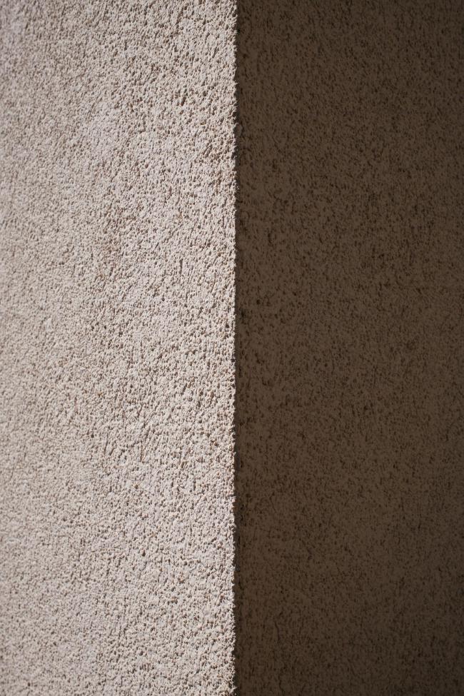 Farbbild im Portrait Format. Blick auf eine raue, verputzte Ecke einer Hauswand. Es gibt keine Referenzpunkte, aber eine Hälfte liegt in der Sonne und eine im Schatten und das Bild wird genau mittig Vertikal dadurch geteilt. Die Farben sind dabei helles und dunkles Gelbgrau.