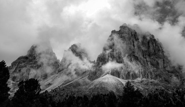 Schwarzweißbild im Landscape Format. Zu sehen ist die Rückseite des Langkofelmassivs aus der Steinernen Stadt heraus. Am unteren Rand sieht man die Spitzen einiger Nadelbäume, dann folgen die Geröllhalten und dann die eigentlichen Gipfel. Die Gipfel sind umrahmt von Wolkenfetzen und verschwinden teilweise auch darin. Der Langkofel rechts im Bild wird auch ein bisschen von der Sonne angestrahlt wodurch insgesamt ein Plastischer und trotzdem mystischer Effekt entsteht.