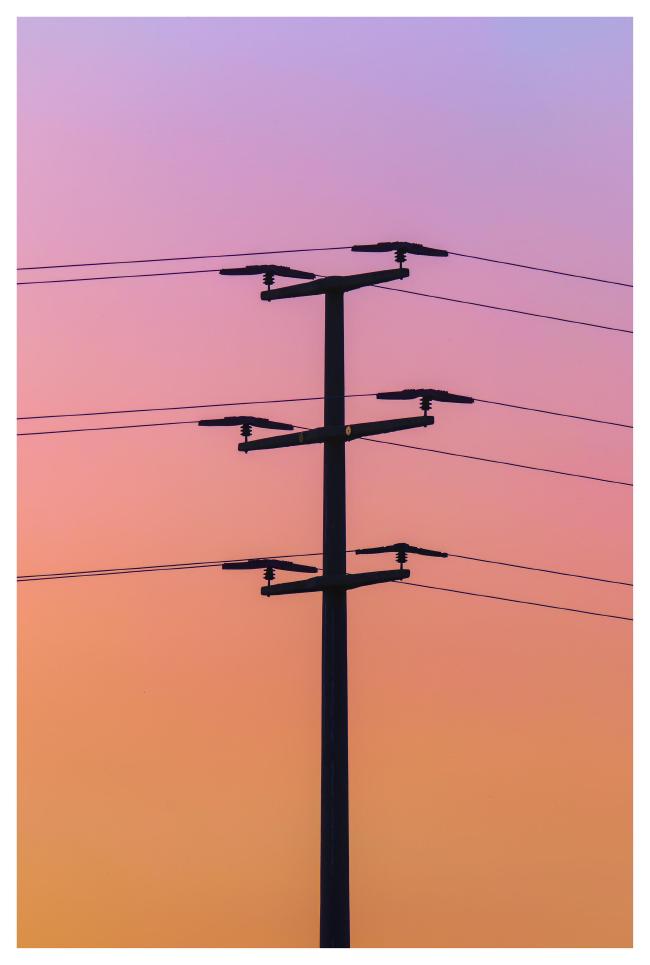 Farbbild im Portrait Format mit weißem Rahmen. Darauf zu sehen ist ein kleiner Strommast mit 6 Leitungen im Profil gegen einen wunderschönen wolkenfreien Himmel mit einem Farbverlauf von oben fliederfarben zu tiefem rot-orange im unteren Bild.