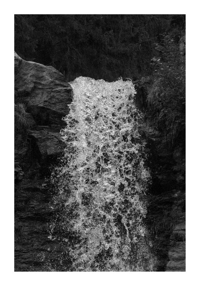 Schwarz-Weiß Bild im Hochkant Format mit weißem Rahmen. Zu sehen ist der selbe Wasserfall wie in Bild 2. Diesmal aber eine Nahaufnahme von der Wasserfallkante und einer kurzen Belichtungszeit. Alles um das Wasser herum ist sehr dunkel nur das Wasser stürzt hell über die Kante.