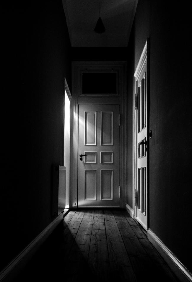 Ein dunkler Flur mit 3 wei0en Türen und holzboden. Aus einem, Raum scheint aus der fast verschlossenen Tür ein heller Lichtschein.