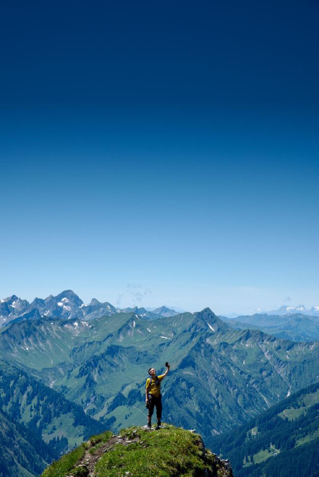 Eine Person mit Rucksacke, gelbem Shirt und schwarzer Hose steht auf einem Felsvorsprung der mit Gras bewachsen ist. Man sieht einen Trampelpfad der zu dieser Stelle führt. Die Person hält ihr Handy hoch um ein Selfie zu machen. Im Hintergrund sind Berger mit wenigen Bäumen aber doch viel Grün zu erkennen. Einige haben noch ein wenig Schnee auf sich. Der Himmel ist fast wolkenlos blau. 