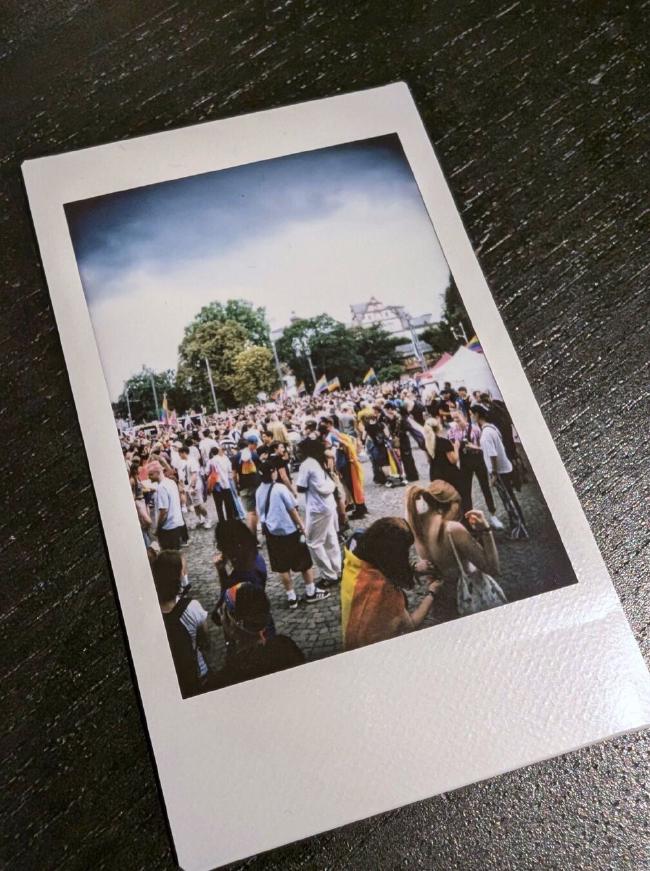 Bild von einem Fujifilm Instax Farbbild auf einer schwarzbraunen Unterlage. Auf dem Bild ist die wartende Menge beim CSD Darmstadt zu sehen.