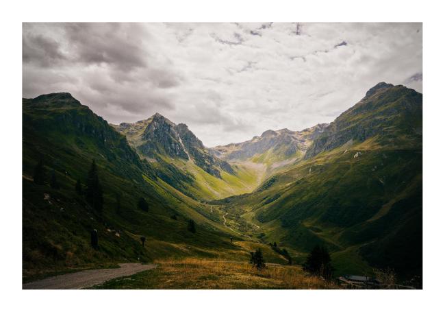 Farbbild im Querformat mit weißem Rahmen. Zusehen ist das grüne sonnen-beschienene Novatal aber diesmal deutlich näher mit einem Forstweg links im Bild. Der Himmel ist inzwischen etwas dunkler und fast flächendeckend Bewölkt und Teile des Tals im vorderen Bild liegen im Schatten. 