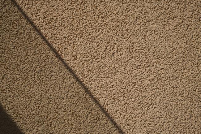 Farbbild im Landscape Format. Zu sehen ist eine raue, gelbe Fassade. Etwas über ein Drittel von Links quert ein diagonaler Schatten von Links oben, nach rechts unten das Bild. Weiter links ist das Bild etwas blauer und dann ist die untere linke Ecke wieder Diagonal durch einen Schatten abgetrennt.