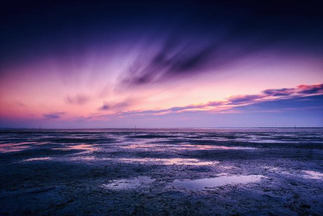 Man sieht das durch Ebbe trockengefallene Watt. In einigen Pfützen spiegelt sich der Himmel. Diese erstrahlt zum Sonnenuntergang in Lila und Blautönen und die Wolken sind durch die Belichtungszeit unscharf 