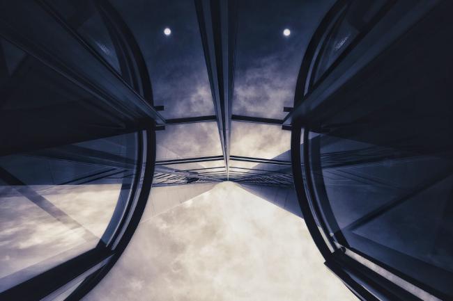 Monochrome Aufnahme. Blick aufwärts an einer verglasten Fassade. Rechts und links sieht man runde Strukturen von den Drehtüren. Der leicht wolkige Himmel spiegelt sich in der Glasfassade und im unteren Teil des Bildes. Im oberen Teil sieht man noch zwei runde Lampen durch die Fassade wodurch das ganze Bild ein wenig wie ein Roboter Gesicht aussieht.