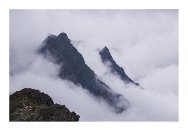 Farbbild im Querformat mit weißem Rahmen. Zu sehen sind zwei große Bergstücke die fast blau wirken zwischen jeder Menge Wolken. Davor in der Linken ecke ist ein näherer Teil Berg auf dem eine Person mit rotem Rucksack und orangener Jacke läuft,