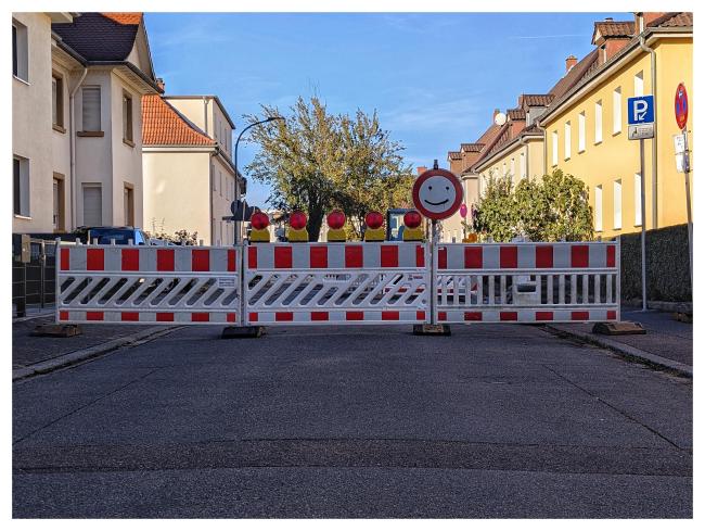 Farbbild mit weißem Rand im Querformat. Zu sehen ist eine Straße die mit Baustellen Absperrungen abgesperrt ist. In der Mitte rote Lichter und daneben ein Durchfahrt Verboten Schild mit einem Smily drin. Dahinter ein paar Häuser und Bäume zum Teil im Schatten zum Teil in der Sonne.