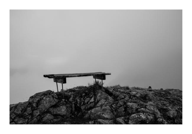 Schwarz-Weiß Bild im Querformat. Zu sehen ist eine rudimentäre Bank auf einer Abbruchkante mit bestimmt wundervoller Aussicht bei klarem Wetter, aber davor verschwimmt alles im Nebel.