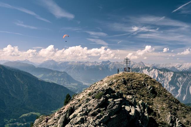 Blick auf einen felsigen, teilweise mit Wiese bewachsenen Berggipfel mit Gipfelkreuz und Einigen Personen m dahin der Blick aus dem Ötztal hinaus. Im Hintergrund sind mehr Berge zu sehen und einige Wolken. Links von der Mitte fliegt ein orangener Paragleiter