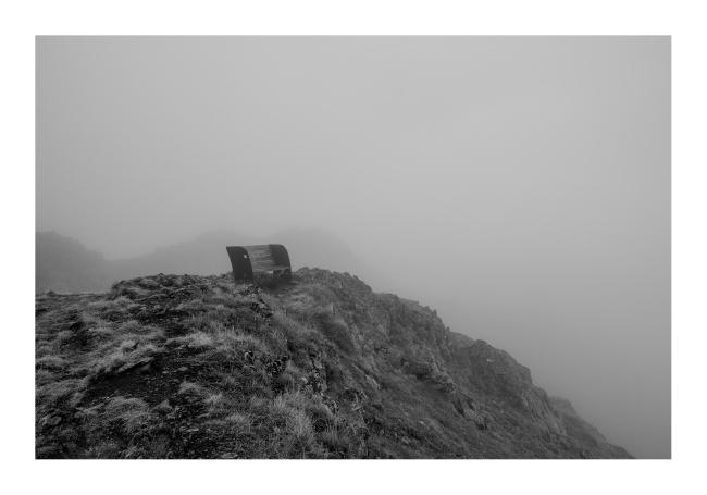 Schwarz-Weiß Bild im Querformat. Zu sehen ist eine Bank auf einer Abbruchkante mit bestimmt wundervoller Aussicht bei klarem Wetter, aber dahinter und davor verschwimmt alles im Nebel.