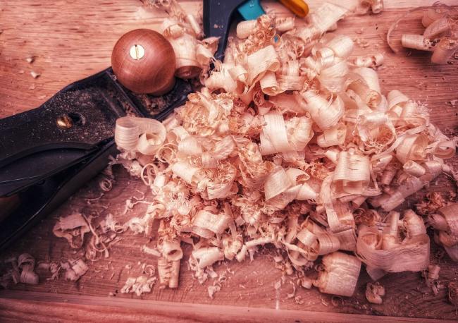 Farbbild Bild in wärmen Tönen und horizontaler Ausrichtung. Den größten Teil des Bildes machen Hobelspäne aus. Links im Bild sieht man einen Teil vom Hobel der weitestgehend schwarz ist mit einem hölzernen Griff. Oben mittig sieht man in schwarz türkis gelb den Griff einer Einhandzwinge