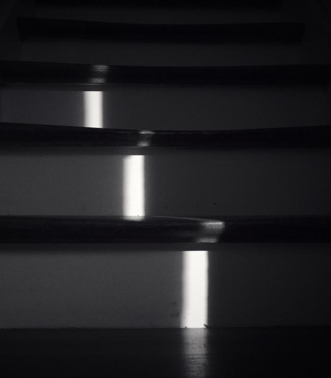 Sehr dunkles, fast quadratisches schwarz-weiß Bild. Zu sehen sind mehrere zum Teil stark abgenutzte Holzstufen, die sehr dunkel sind. Der Raum zwischen den Stufen ist weiß angemalt. Von unten leicht rechts der Mitte nach oben Links der Mitte zieht sich ein Lichtstreifen der fast nur auf den weißen Bereichen wiederum stufig zu sehen ist.