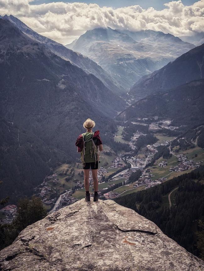 Farbfoto im Portrait Format. Eine Person (von hinten fotografiert) mit Strohhut, rotem T-Shirt, grünem Rucksack kurzer schwarzer Hose und Wanderschuhen steht an der Spitze eines dreieckig wirkenden Felsens in der mitte der unteren Hälfte des Bildes. Der Fels macht ungefähr ein Drittel des Bildes aus. Es wirkt als stände der Felsvorsprung weit über dem Abgrund. Im Hintergrund ist eine Dorf im Tal zu sehen sowie Bergflanken rechts und links davon. Im Tal Schluss ist auch noch ein Berg zu sehen der etwas mehr Sonne abbekommt als die rechts und links. Über dem hinteren Berg hängen Wolken direkt über dem Gipfel und es ist leicht diesig im Tal. Über den Wolken sieht man noch etwas blauen Himmel.