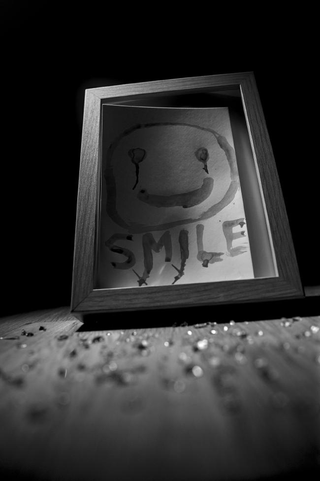 Schwarzweißfoto im Portrait-Format. Ein Bilderrahmen steht leicht schief auf einer Holzoberfläche und beides wird von Links oben angestrahlt. Das Licht hört hinter dem Bilderrahmen aber sofort auf und damit liegt der ganze Hintergrund im Dunkeln. Im Bilderrahmen ist ein Stück weißes Papier in Postkartengröße. Darauf zu sehen ist ein Smily unter dem steht in Großbuchstaben SMILE. Alles sieht so aus als wäre es grob mit einem Finger gezeichnet bzw. geschrieben. Der Smily liegt dabei im Halbschatten der sich leicht aus dem Rahmen wölbenden Karte da der Rahmen keine Scheibe mehr hat. Sowohl die Augen des Smilies als auch die Buchstaben M und I  verlaufen nach unten wodurch es so aussieht als würde der Smily lachend weinen. Alles hat eine leicht unangenehme Stimmung, besonders wenn man weiß dass es mit Blut gemalt ist.  Hier liegen zusätzlich zum vorherigen Bild noch kleine Glasscherben vor dem Bilderrahmen und das Bild ist von unterhalb des Rahmens aufgenommen und das ganze Bild ist leicht nach links gekippt, was das unangenehme Gefühl nochmal verstärkt, dass etwas aus der Balance geraten ist.