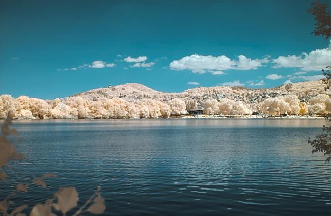 Farbbild im Landscape Format. Links und Rechts ragen ein paar zweige ins Bild. Dahinter liegt der blaue See und im Hintergrund folgen dann das Strandbad und Bäume um den See. Außerdem die Ausläufer des Odenwalds. Die Bäume dort sind alle eher Gelbweiß und der Himmel ist azurblau mit ein paar wenigen Wolken.
