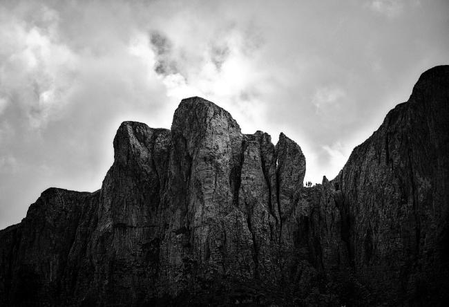 Schwarz-Weiß Bild: Der Himmel ist bewölkt und macht etwa 1/3 des Bildes aus. Darunter sieht man dunkel aber stark strukturiert eine Bergflanke die in der Mitte heller Beleuchtet ist. Rechts von der Mitte gibt es eine kleine Scharte in der drei Menschen stehen.