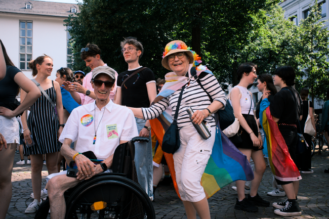 Farbbild, zwei ältere Menschen lachen in die Kamera und haben dabei verschiedene Aufkleber, Flaggen oder ähnliches in Regenbogenfarben am Körper. Der (vermutlich) Mann sitzt im Rollstuhl und die (ebenso vermutlich) Frau hat eine Hand auf seiner Schulter. Im Hintergrund sind noch ein paar Personen in Grüppchen die sich unterhalten und ein paar Bäume und Gebäude.