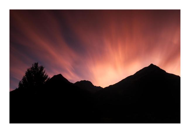 Farbbild im Querformat. Hinter einer schwarzen Bergkulisse sieht man die durch Langzeitbelichtung verschwommenen rot und golden angeleuchteten Wolken eines Sonnenuntergangs. 