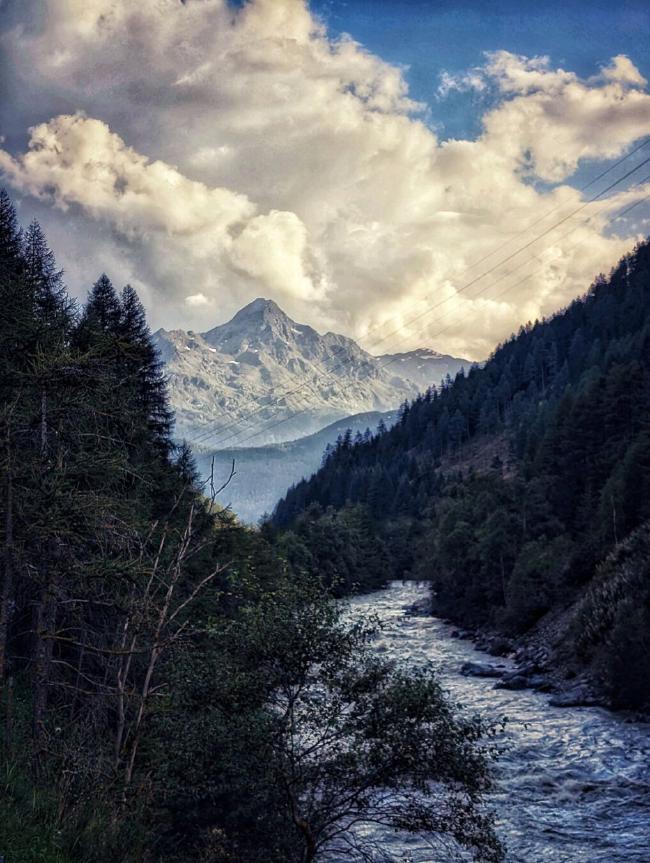 Blick über einen Gebirgsfluss gesäumt von Nadelbäumen der sich in den Hintergrund schlängelt. Im Hintergrund ein majestätischer schroffer Berg und darüber kontrastreiche von der untergehenden Sonne beleuchtete Wolken