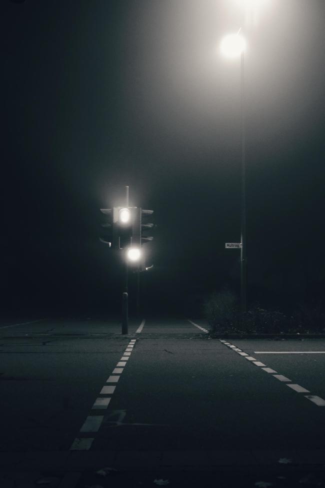 Eine schwarz-weiß Bild im Portrait Format. Man sieht die Markierungen für einen Ampel Übergang leicht rechts der Mitte. Bei der Fußgänger Ampel leuchtet das obere Licht. Im rechten Bildteil steht eine Laterne die einen Runden Halo im Nebel wirft der auch das licht der Ampel streut. Auf der anderen Straßenseite wartet aber nur Dunkelheit auf einen.