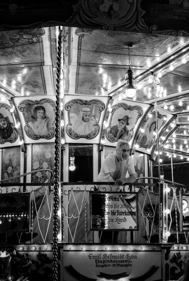 Schwarzweißbild im Portrait Format. Ein älterer Mann mit weißem T-Shirt steht auf der oberen Plattform eine alten Etagenkarusells dem man seine Jahre schon ansieht. Überall sind leuchtende Birnen zu sehen. Der Mann lehnt dabei auf dem Geländer mit einer offenen Hand am Gesicht und wirkt unglaublich erschöpft. Der Kopf ist zwar leicht von der Kamera weg ausgerichtet, aber die Augen schauen in Richtung der Kamera.