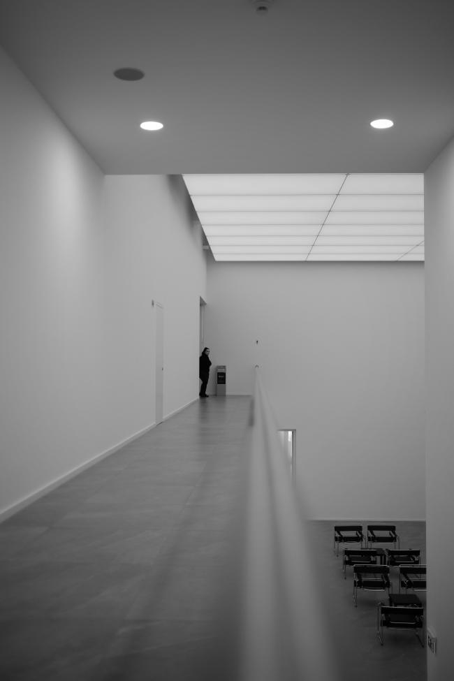 Schwarzweißbild im Portrait Format. Die Bildmitte wird von einer unscharfen Edelstahlbrüstung dominiert die ich bis in die Bildmitte zieht. Auf der rechten Seite sieht man einen tiefer gelegenen Bereich mit einigen Stühlen. Im linken Bereich sieht man einen Gang an dessen Ende eine Person steht die in Richtung der Kamera guckt. Die Person ist ganz in Schwarz Gekleidet. Das obere Drittel macht eine Decke mit Punktstrahlern aus, etwas dahinter liegt eine weitere höhere Decke mit Raumfüllenden fast randlosen Licht Paneelen. Alles wirkt sehr clean da alle Wände leer sind und auch in den Räumen bis auf die Stühle nichts steht.