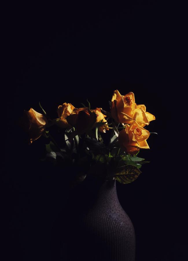 Farbfoto im Portrait Format. Man sieht einen Strauß gelber Rosen die schon bessere Zeiten hatten in einer grauen grillten Vase vor einem schwarzen Hintergrund. Die Blumen sind von Links angeleuchtet. Das Bild ist insgesamt low-key.