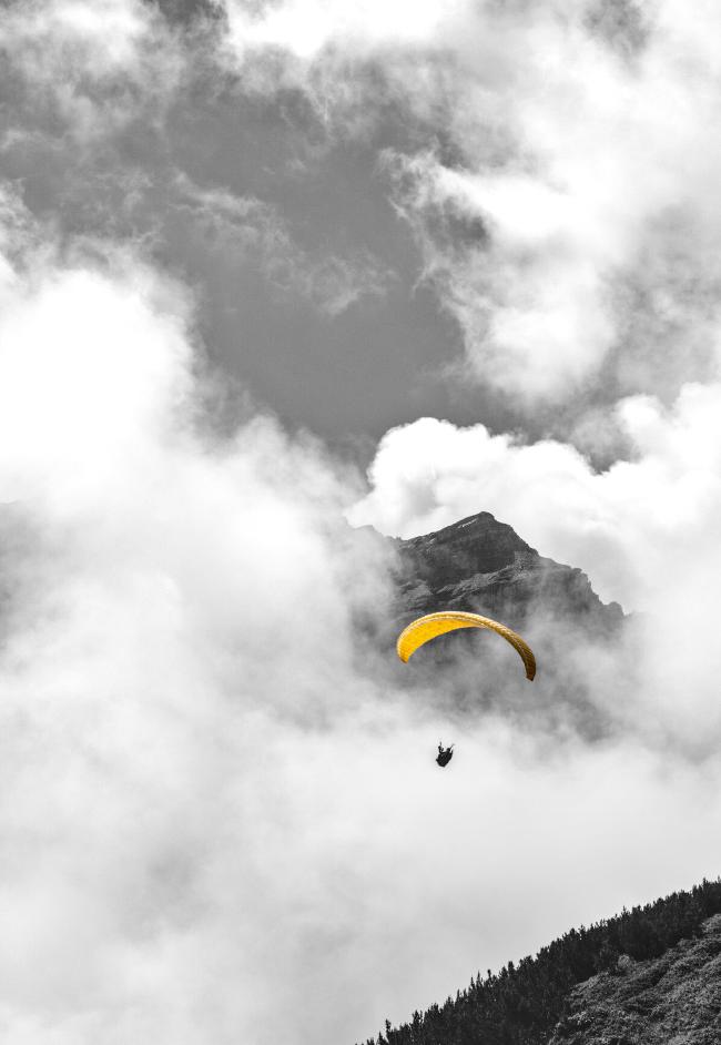 Paraglider umgeben von Wolken. Im Hintergrund ist eine Bergspitze zu erkennen. Das Bild ist Schwarz-Weiß nur der Schirm des Paragliders leuchtet in Gelb.