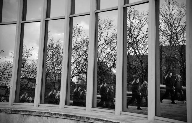 Blick auf eine stark spiegelnde leicht gebogen verlaufende Fensterfront. In dem Fenster sieht man einen unterschiedlichen Ausschnitt von zwei Personen, einem Baum und einem Gebäude im Hintergrund.