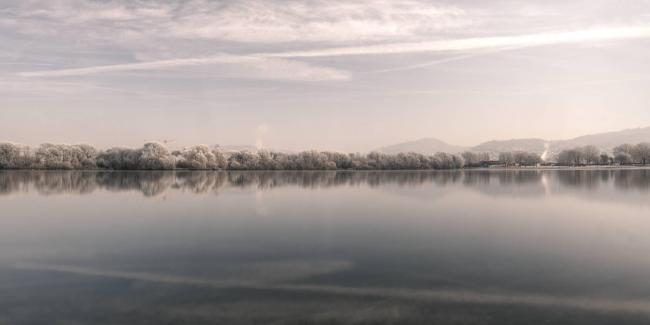Farbbild im Landscape Format. Man sieht den Blick über einen See in dessen glatter Oberfläche sich die Wolken spiegeln. In der Bildmitte liegt das andere Ufer mit weiß überzogenen Bäumen. Auf der rechten Seite sieht man ein paar Berge der Bergstraße