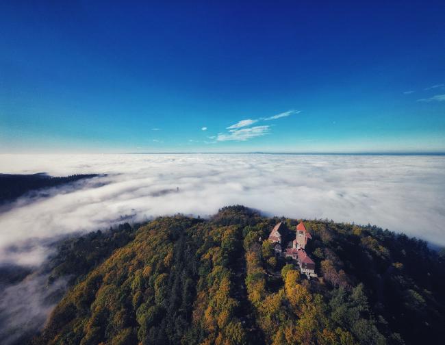 Luftaufnahme einer Drohne die über einem herbstlichen Wald am Rande einer Mittelgebirgskette fliegt. Der Blick geht in die von Nebel geflutete Ebene. In der Bild Mitte schaut eine Burg aus dem Nebel. Etwas weiter hinten eine Zweite. Der Himmel ist strahlend Blau.