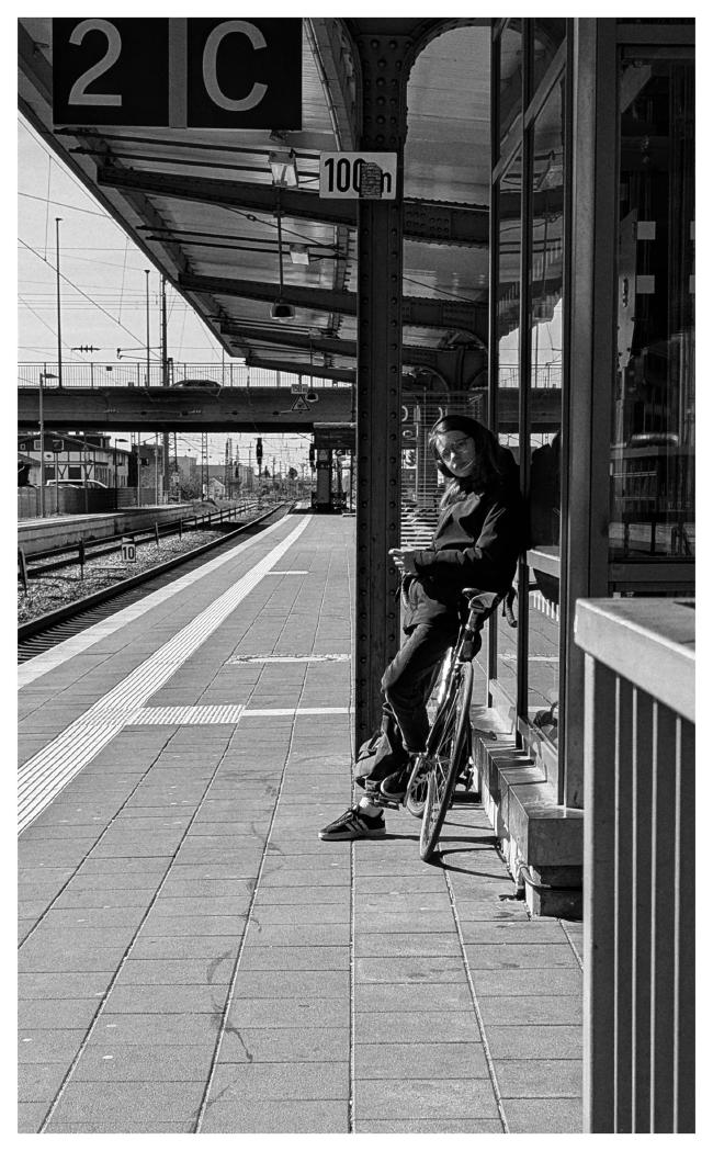 Schwarzweiß Bild im Portrait Format mit weißem Rahmen. Eine Person seht auf einem überdachten Bahnsteig an ihr Fahrrad angelehnt vor einem Fahrstuhl. Im Hintergrund führt eine Brücke über die Gleise und den Bahnsteig. Die Person guck in Richtung der Kamera aber nicht direkt sondern auf den Abfahrtsanzeiger dahinter.