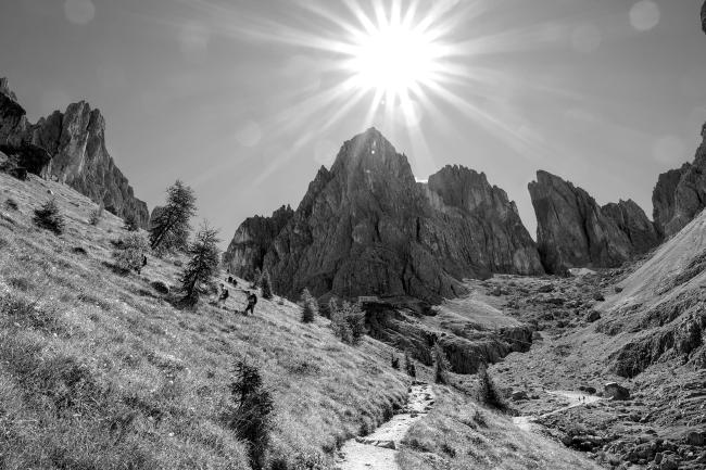 Schwarz-Weiß Bild im Landscape Format. Man sieht einen Talkessel umrandet von schroffen Berggipfeln. Das Tal selbst ist eher grassig mit ein paar kleinen Bäumen. Über dem Gipfel in der Mitte steht die Sonne die einen vielzackingen Stern wirft und ein paar kleine Bokeh Punkte hinterlässt. In der Mitte des Bildes sieht man einen Weg der sich dann an der linken Bildseite hochschlängelt. Dort sieht man ein paar weitere Wandernde. Etwas Rechts der Mitte sieht man noch einen Versorgungsweg. und in der Bildmitte unter dem höchsten Gipfel ist die Hütte zu sehen.