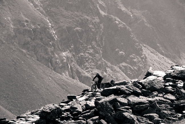 Schwarz Weiß Bild einer Person die mit dem Mountainbike über Felsplatten eine Berg hinab fährt. Im Hintergrund ist der Hang eines weiteren Berges zu erkennen.