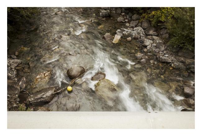 Farbbild im Querformat mit weißem Rahmen. Zu sehen ist ein Flussbett, das der Ill, in dem das Wasser eine leichte Bewegungsunschärfe hat. Mir hat es da besonders der gelbe Ball angetan der im unteren linken Drittel blockiert von einem Ast in einem ruhigeren Bereich des Flusses schwimmt.