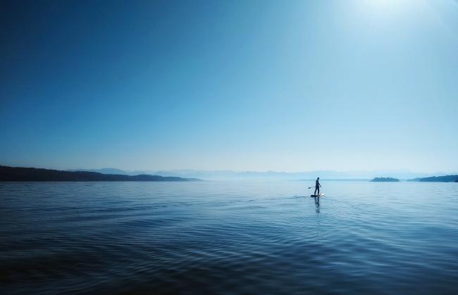 Ein Stand-Up-Paddler auf einem See. Im Hintergrund sind im Diesigen Berge zu erkennen. Es ist strahlender Sonnenschein und das Bild ist in Blautönen gehalten.