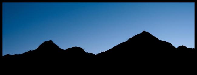 Farbbild im Ultrabreit-Format (XPan) mit schwarzem Rahmen. Zu sehen ist die Gipfelsilhouette einer Bergkette mit ein paar Bäumen hier und da auf der Kamlinie. Dahinter der nahezu wolkenfreie Himmel in einem tiefen Blau links und einem etwas helleren Blau rechts.
