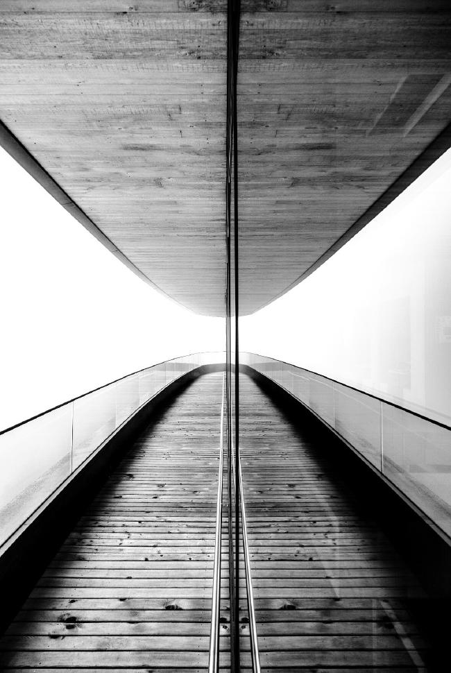Schwarz-Weiß Bild. Auf dem Bild sieht man eine Terasse aus Holz mit einem Geländer aus Glas. Darüber eine Decke aus Holz. Nach hinten macht das ganze einen Bogen. In der Mitte Reflektiert sich die Szene an einer Glasfront wodurch der Effekt einer Parabel entsteht. An der Galsfront ist noch ein Handlauf aus Stahl angebracht. Im hinteren Teil des Bildes wird es etwas heller. Durch den dichten Nebel ist keine Umgebung erkennbar.