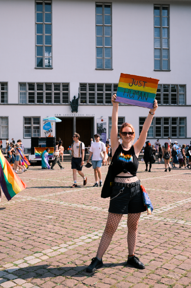 Farbbild einer Person mit einem ärmellosen und bauchfreien Top einer schwarzen kurzen Hose und einer Netzstrumpfhose steht auf einem Platz vor einen Gebäude und hält ein Schild in Regenbogenfarben mit dem Text Just Human hoch. Im Hintergrund laufen noch weitere Personen über den Platz vorm Gebäude.