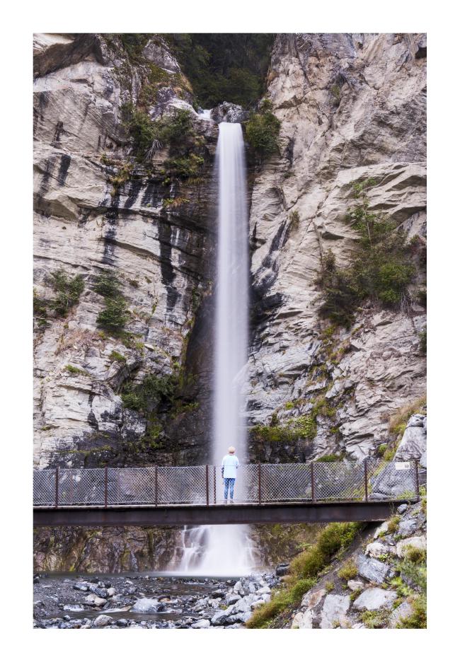 Farbbild im Hochkant Format mit weißem Rahmen. Zu sehen ist ein ziemlich gerader Wasserfall gerahmt von hellem Gestein und ein paar Bäumen hier und da. Hinter dem Wasserfall ist der Stein schwarz bzw. sehr dunkel gefärbt. Davor ist eine Brücke auf der genau vor dem Wasserfall eine Person mit hellgrünem Oberteil und blauer Jeans steht. Der Wasserfall hat Bewgungsunschärfe durch eine längere Belichtungszeit.