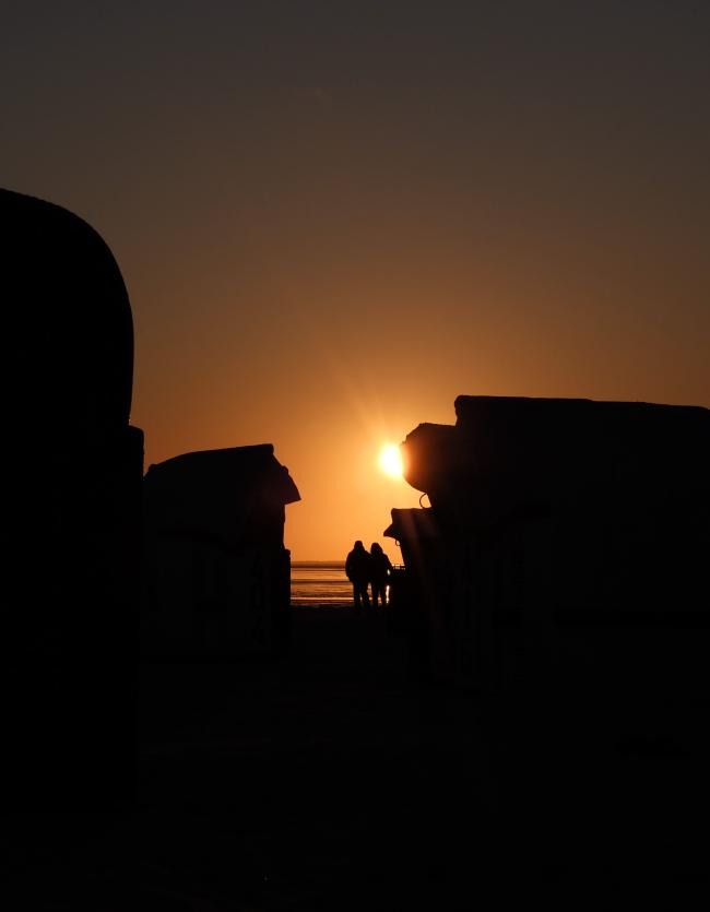 Farbbild im Portrait Format. Zwei Personen laufen sehr nah beieinander in Richtung Sonnenuntergang. Die Szene ist gerahmt durch mehrere Strandkörbe die wie die Personen aber nur als schwarze Silhouette erkennbar sind. Vor den Menschen ist ein wenig Meer zu erkennen. Über den Menschen, leicht verdeckt durch einen Strandkorb sieht man die untergehende Sonne. Alles ist in ein sanftes orangenes Licht getaucht.