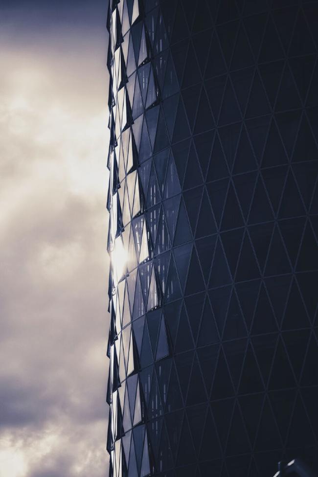 Bild in Portrait Orientierung von einer Hochhausfront aus dreieckigen Scheiben. Einige davon stehen wie Schuppen leicht geöffnet hervor. Die Sonne spiegelt sich in einigen der Scheiben und der Himmel wirkt leicht düster. Das Bild relativ monochrom allerdings gehen die Farben leicht ind gelb/blau.