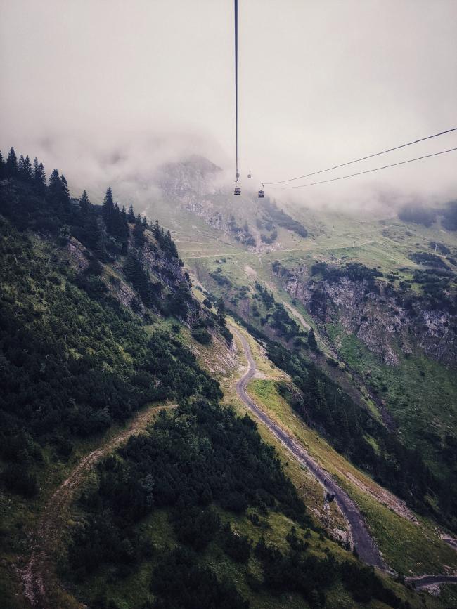 Man sieht eine Berglandschaft fotografiert aus einer Gondel. Auf den Hängen stehen einige Bäume und ein Forstweg führt nach oben. Am oberen Bildrand liegt der Berg in den Wolken und Seile sowie Gondeln führen dort hin.