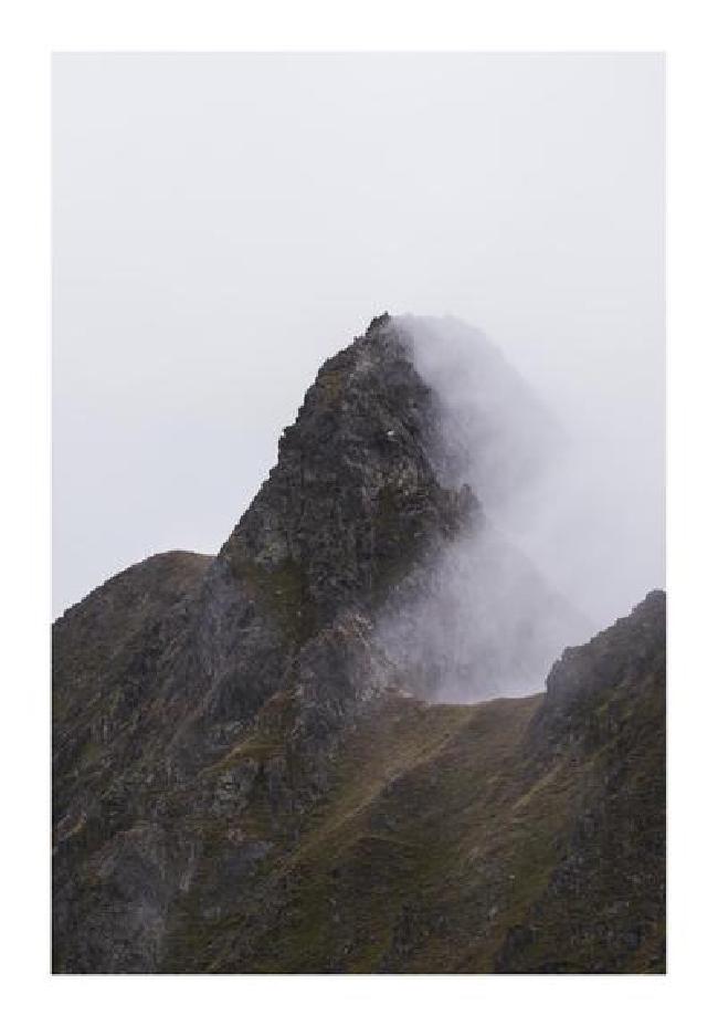 Farbbild im Porträt Format mit weißem Rahmen. Das Bild zeigt einen Berggipfel an einem Kamm  Zu sehen ist wie der Nebel auf der einen Seite vom Kam am Berg hängt, während die andere Seite noch frei sichtbar ist.