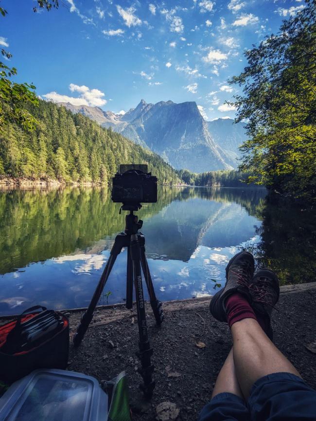 You see my legs and camera on a tripod infront of a lake with a beautiful mountain landscape behind it. It is sunny and there are only a few clouds on the blue sky